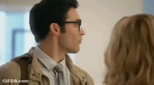 A Man Wearing Glasses And A Tan Jacket Is Talking To A Woman In A Room . GIF