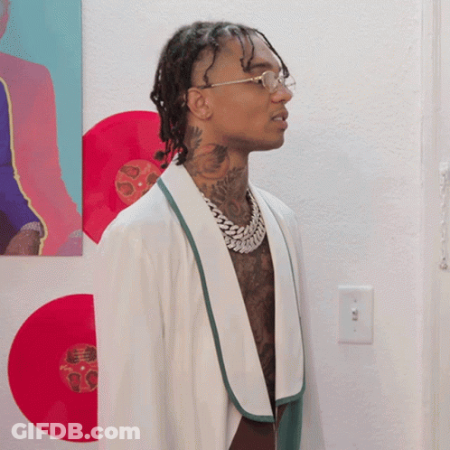 A Man Wearing Glasses And A White Robe Is Standing In Front Of A Wall With Red Records On It GIF