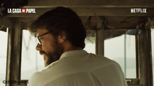 A Man Wearing Glasses And A White Shirt Is Sitting In Front Of A Sign That Says La Casa De Papel And Netflix GIF