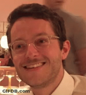A Man Wearing Glasses And A White Shirt Is Smiling And Looking At The Camera . GIF