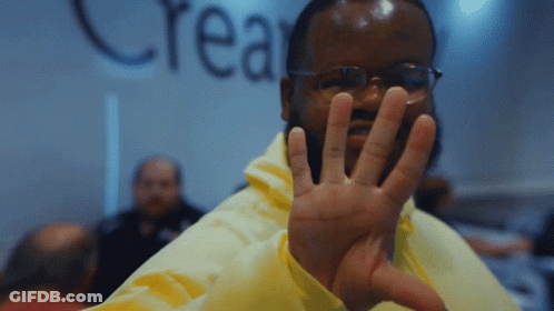 A Man Wearing Glasses And A Yellow Jacket Holds Up His Hand In Front Of A Wall That Says Crea GIF
