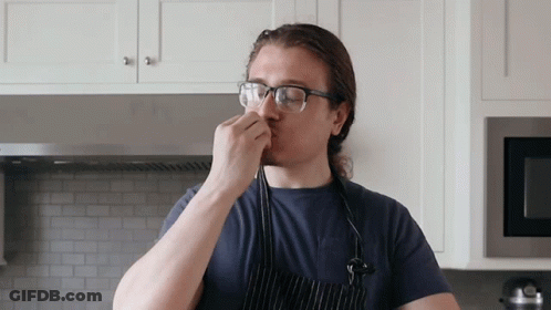 A Man Wearing Glasses And An Apron Smells Something In A Kitchen GIF