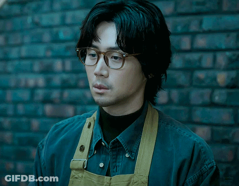 A Man Wearing Glasses And An Apron Stands In Front Of A Black Brick Wall GIF