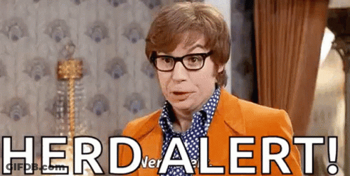 A Man Wearing Glasses And An Orange Suit Is Standing In A Room With The Words Herd Alert Written On The Wall . GIF