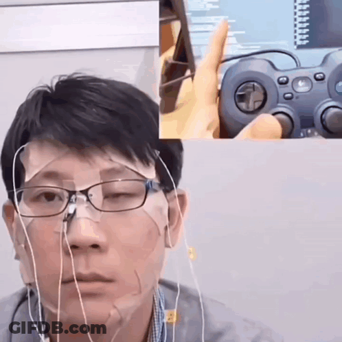 A Man Wearing Glasses And Earbuds Is Holding A Logitech Controller In Front Of His Face GIF