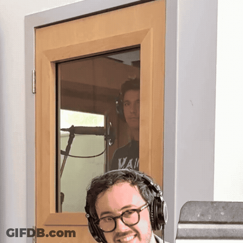 A Man Wearing Glasses And Headphones Stands In Front Of A Door That Has A Window GIF