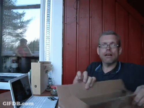 A Man Wearing Glasses Holds A Cardboard Box In Front Of A Laptop Computer GIF
