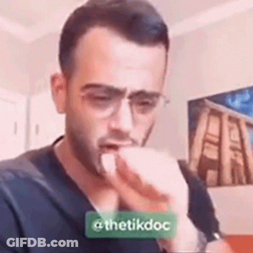 A Man Wearing Glasses Is Coughing Into His Hand In A Living Room . GIF