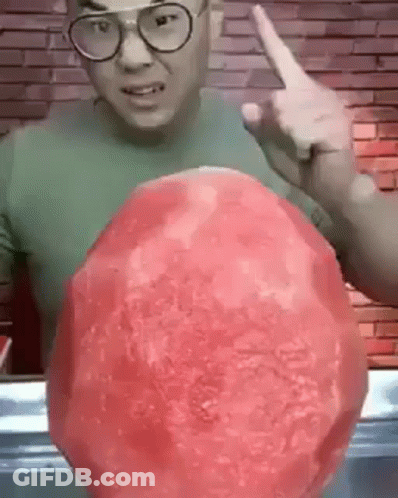 A Man Wearing Glasses Is Holding A Large Watermelon In His Hand And Pointing At It . GIF