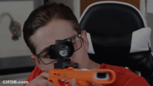 A Man Wearing Glasses Is Holding A Nerf Gun In Front Of His Face GIF