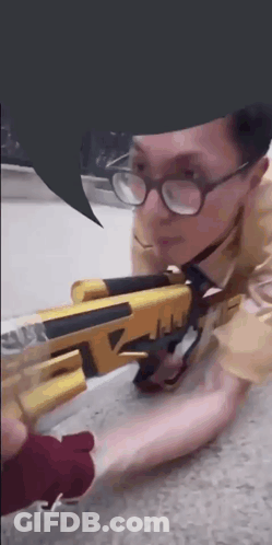 A Man Wearing Glasses Is Kneeling Down With A Gun GIF