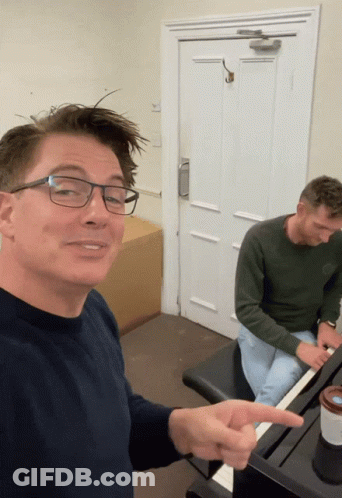 A Man Wearing Glasses Is Pointing To Another Man Playing A Piano GIF
