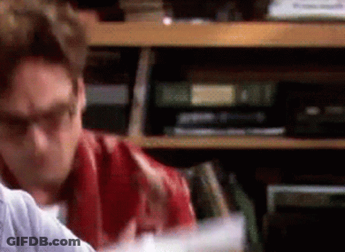 A Man Wearing Glasses Is Sitting In Front Of A Bookshelf Filled With Books GIF