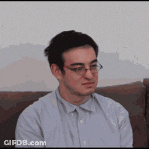 A Man Wearing Glasses Is Sitting On A Couch And Looking At The Camera . GIF