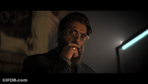 A Man Wearing Glasses Is Smoking A Cigarette In Front Of A Wall With Arabic Writing On It GIF