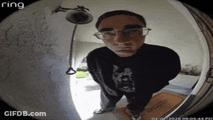 A Man Wearing Glasses Is Standing In Front Of A Doorbell Looking At The Camera . GIF