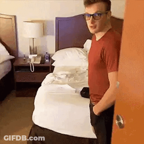 A Man Wearing Glasses Is Standing In Front Of A Hotel Bed . GIF
