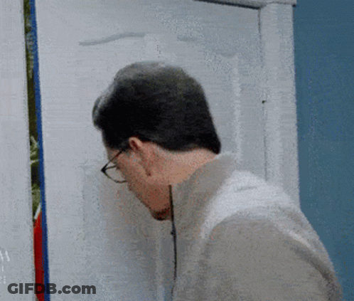 A Man Wearing Glasses Looks Through A Hole In A Door GIF