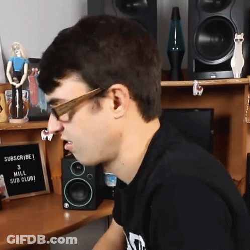 A Man Wearing Glasses Sits In Front Of A Sign That Says " Subscribe 3 Mill Sub Club " GIF