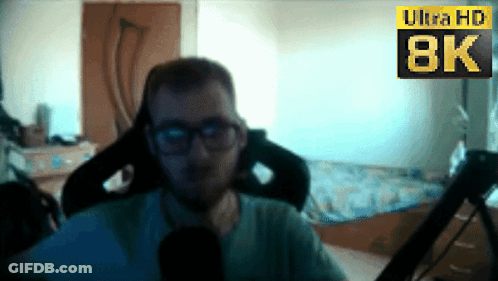 A Man Wearing Glasses Sits In Front Of An 8K Sign GIF