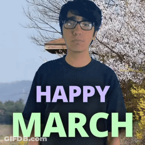 A Man Wearing Glasses Stands In Front Of A Sign That Reads Happy March GIF