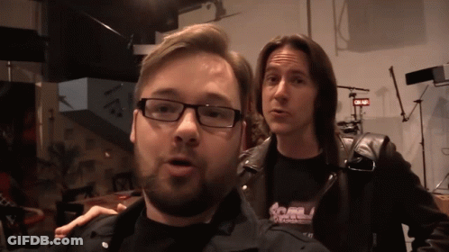 A Man Wearing Glasses Stands Next To Another Man Wearing A Black Shirt That Says ' S.a.m. ' GIF