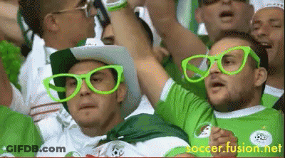 A Man Wearing Green Glasses And A Cowboy Hat Stands In A Crowd Of People GIF