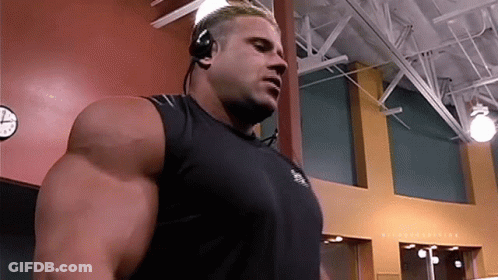 A Man Wearing Headphones And A Black Tank Top Stands In Front Of A Clock GIF