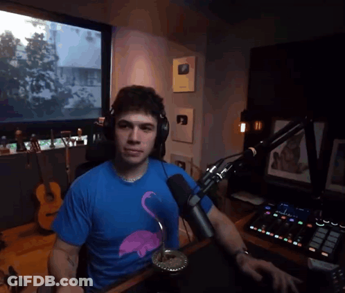 A Man Wearing Headphones And A Blue Flamingo Shirt Is Sitting At A Desk In Front Of A Microphone GIF