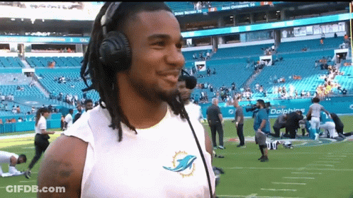 A Man Wearing Headphones And A Dolphins Shirt Is Standing On A Football Field GIF