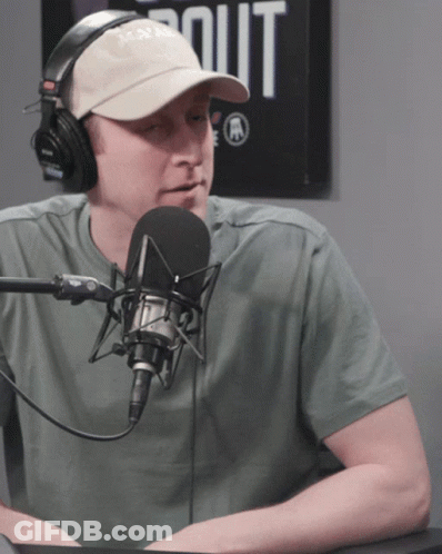 A Man Wearing Headphones And A Hat Talks Into A Microphone In Front Of A Sign That Says Out GIF