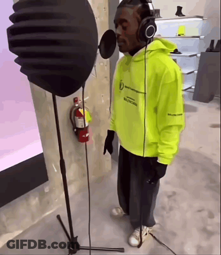 A Man Wearing Headphones And A Neon Yellow Hoodie Sings Into A Microphone GIF