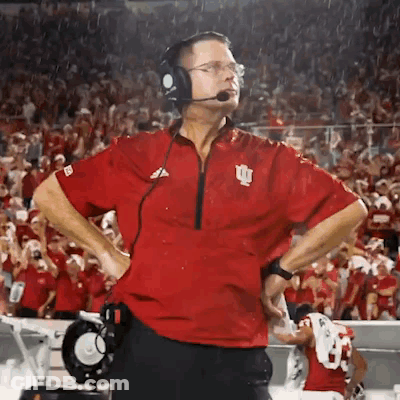 A Man Wearing Headphones And A Red Shirt With The Letter I On It Stands On A Football Field . GIF