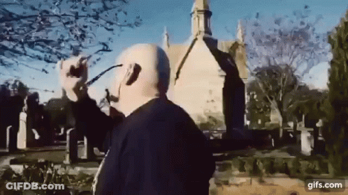 A Man Wearing Headphones And Glasses Is Standing In Front Of A Church In A Cemetery . GIF
