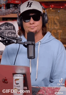 A Man Wearing Headphones And Sunglasses Is Sitting In Front Of A Microphone And A Laptop . GIF