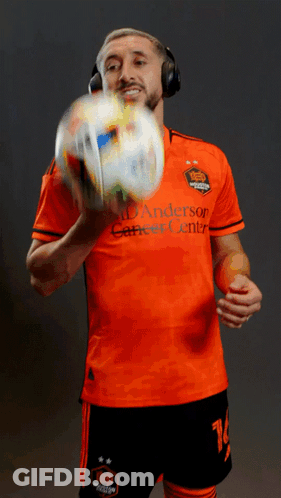 A Man Wearing Headphones Holds A Soccer Ball While Wearing An Orange Anderson Career Center Jersey GIF