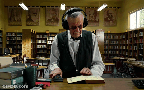 A Man Wearing Headphones Is Reading A Book In A Library With Posters Of Darwin And Albert Einstein On The Wall GIF