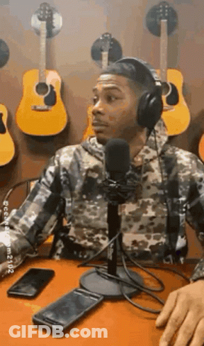 A Man Wearing Headphones Is Sitting At A Table In Front Of A Microphone In Front Of A Wall Of Guitars . GIF