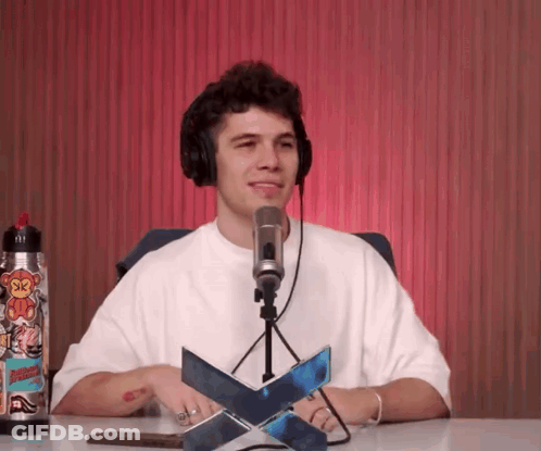 A Man Wearing Headphones Is Sitting In Front Of A Microphone With A Sticker On His Water Bottle That Says Sk GIF