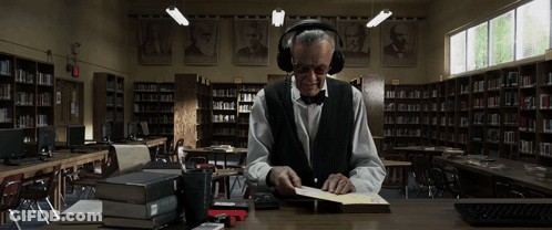 A Man Wearing Headphones Sits At A Desk In A Library Reading A Book GIF