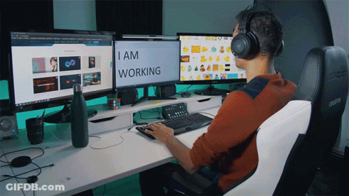 A Man Wearing Headphones Sits At A Desk With Three Computer Monitors And The Words I Am Working On The Screen GIF