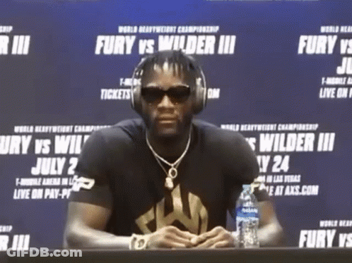 A Man Wearing Headphones Sits At A Table With A Bottle Of Water In Front Of A Sign That Says Fury GIF