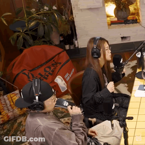 A Man Wearing Headphones Sits Next To A Woman Wearing Headphones In Front Of A Red Bag That Says Lion GIF