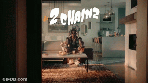 A Man Wearing Headphones Sits On A Couch In A Living Room With 2 Chainz Written On The Wall Behind Him GIF