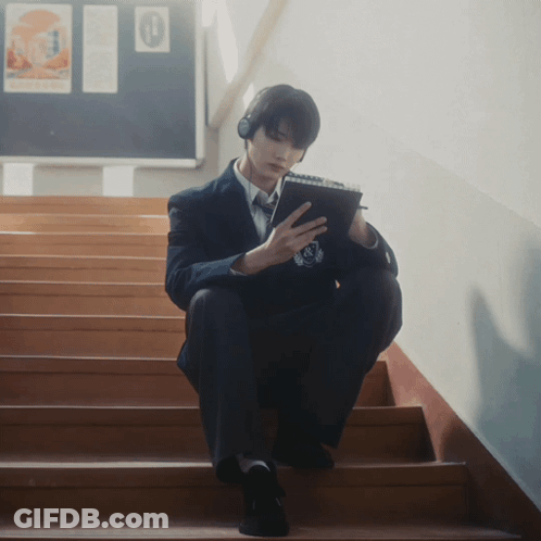 A Man Wearing Headphones Sits On A Set Of Stairs Looking At His Phone GIF