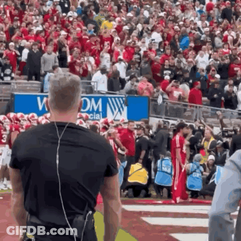 A Man Wearing Headphones Stands In Front Of A Crowd At A Football Game .. GIF