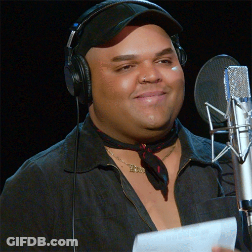 A Man Wearing Headphones Stands In Front Of A Microphone With A Necklace That Says Exi GIF