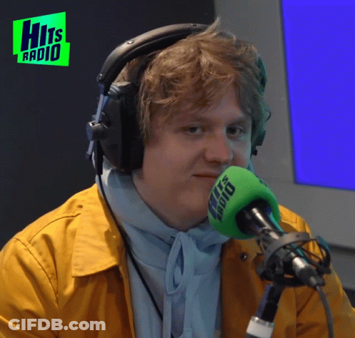 A Man Wearing Headphones Stands In Front Of A Microphone With The Words Hits Radio In The Background GIF