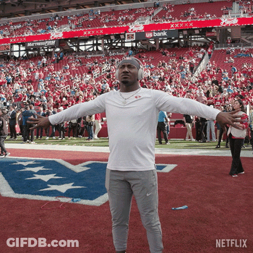A Man Wearing Headphones Stands On A Football Field With His Arms Outstretched In Front Of A Us Bank Banner GIF