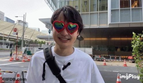 A Man Wearing Heart Shaped Sunglasses Is Smiling In Front Of A Large Building GIF